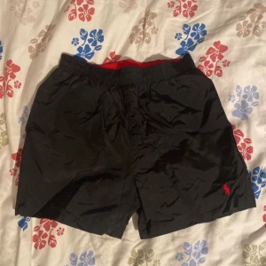 Svarta shorts från Polo Ralph Lauren - Snygga svarta shorts från Polo Ralph Lauren med röd broderad logga. De har en elastisk midja med röd insida och är perfekta för en avslappnad stil. Materialet är lätt och bekvämt. Helt oanvända och lappen på deras ordinarie pris sitter kvar