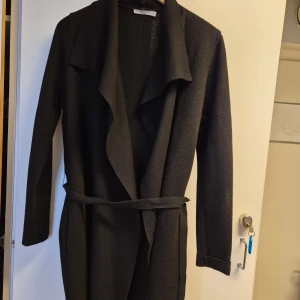 Svart kappa från Wera - Elegant svart kappa från Wera med långärmad design och ett knytbälte i midjan. Perfekt för en stilren look under kyligare dagar.