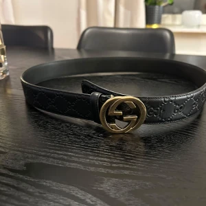 Svart bälte med guldspänne från Gucci - Snyggt svart bälte i skinn från Gucci med ett elegant guldspänne. Bältet har ett diskret mönster som ger det en lyxig känsla. Perfekt för att lyfta vilken outfit som helst. Nypris 4800kr.