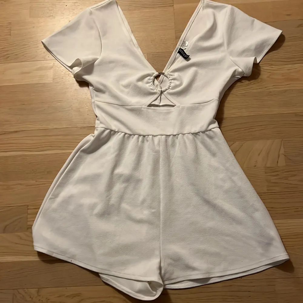 Snygg vit kort jumpsuit från H&M med v-ringning och knytning framtill. Den har korta ärmar och en elastisk midja för en bekväm passform. Perfekt för varma dagar!. Mekot.