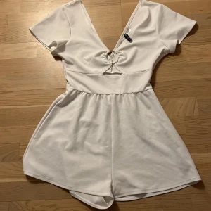 Vit kort jumpsuit från H&M - Snygg vit kort jumpsuit från H&M med v-ringning och knytning framtill. Den har korta ärmar och en elastisk midja för en bekväm passform. Perfekt för varma dagar!