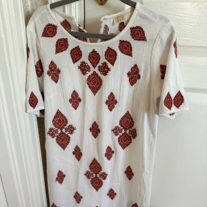 Vit klänning med rött mönster från Michael Kors - Charmig vit klänning med röda broderade mönster. Den har en rund hals och korta ärmar, perfekt för en somrig look. Klänningen är i ett lätt och luftigt material.