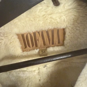 Brun skinnjacka från Jofama - Säljer en snygg brun skinnjacka från Jofama med mjukt foder och krage i pälsimitation. Jackan har en dragkedja och spänne vid kragen för extra stil och värme. Perfekt för kyliga dagar!