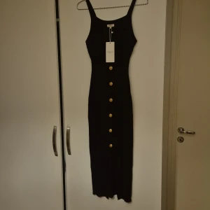 Svart ribbad klänning från raglady  - Snygg svart ribbad klänning från Moccou inköpt på raglady med guldknappar framtill. Klänningen har en tight passform och är ärmlös, perfekt för en stilren look. Den är gjord i ett mjukt material som ger en bekväm känsla.