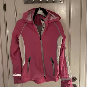 Rosa regnjacka från Daily Sports - Snygg rosa regnjacka från Daily Sports med dragkedja och huva. Jackan har vita och grå detaljer samt praktiska fickor med dragkedjor. Perfekt för regniga dagar.