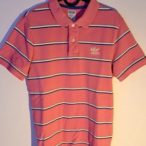 Tommy Hilfiger Denim piké - Snygg rosa pikétröja från Hilfiger Denim med vita och blå ränder. Tröjan har korta ärmar och en klassisk krage med knappar. Perfekt för en avslappnad stil.