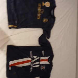 Fotbollströjor från Real Madrid och PSG - Två snygga fotbollströjor: en Real Madrid-tröja i mörkblått med guldtryck från Adidas och en PSG-tröja i mörkblått med röd och vit rand från Nike. Perfekt för fotbollsfans som vill visa sitt stöd för sina favoritlag.