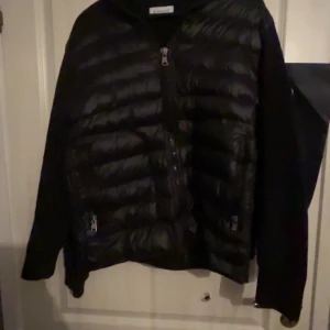 Svart dunjacka från Moncler - Säljer en snygg svart cardigan från Moncler med dragkedja och två fickor framtill. Jackan har en klassisk design med långärmade ärmar och Moncler-logga på armen. Perfekt för kyligare dagar.