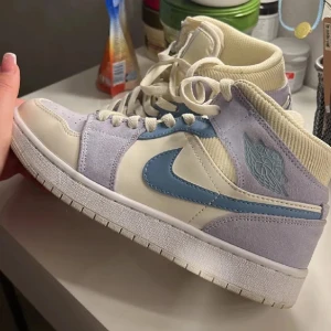 Air Jordan 1 Mid Mixed Textures Blue Tan - Snygga Air Jordan 1 Mid sneakers i en mix av blå och beige nyanser. Skorna har en stilren design med snörning och en ikonisk Nike-logga på sidan. Perfekta för dig som vill ha en sportig och trendig look. (Använda max 2 ggr)