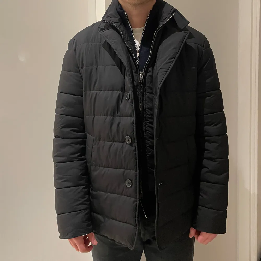 Snygg svart quiltad jacka från John Henric med avtagbar huva. Jackan har både dragkedja och knappar framtill. Inbyggd väst som är avtagbar.. Takit.