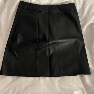 Svart kjol från Bik Bok - Snygg svart kjol i skinnimitation från Bik Bok. Kjolen har en stilren design med en liten slits på sidan och en dragkedja i sidan. 