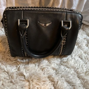 Svart handväska med dragkedja - Snygg svart handväska i skinn med silverdetaljer och dragkedja. Väskan har ett stilrent emblem framtill och två robusta handtag. Perfekt för att bära dina dagliga nödvändigheter med stil.