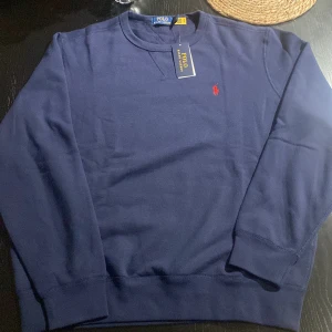 Mörkblå tröja från Polo Ralph Lauren - Snygg mörkblå tröja från Polo Ralph Lauren med det ikoniska röda logotypbroderiet på bröstet. Tröjan har en klassisk rund halsringning och långa ärmar. Perfekt för en stilren och avslappnad look.ny pris på den är 2 tusen som ni ser på sista bilden o tröjan är helt ny fick den i julklapp så har ni funderingar eller liknande är de bara att höra av sig mvh nino