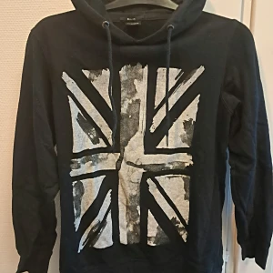 Svart hoodie med tryck från FSBN - Säljer en svart hoodie från FSBN med ett stort tryck av en flagga i grått och vitt på framsidan. Tröjan har en justerbar huva med dragsko och långa ärmar. Perfekt för en casual look!