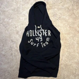 Svart hoodie från Hollister - Säljer en svart hoodie från Hollister med texten 'Hollister 49 Surf Team' på framsidan. Den har en klassisk känguruficka och justerbar huva med snören. Perfekt för en avslappnad stil. Säljer pga för liten för mig. Hoodien är riktigt skön och snygg.