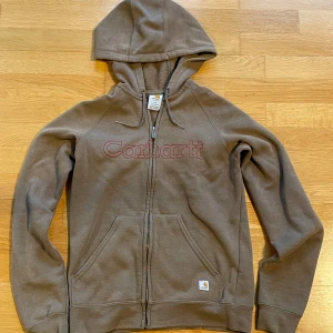 Brun hoodie från Carhartt - Säljer en brun hoodie från Carhartt med dragkedja och broderad logga i rött på framsidan. Tröjan har en mysig huva och praktiska fickor framtill. Perfekt för en avslappnad stil.