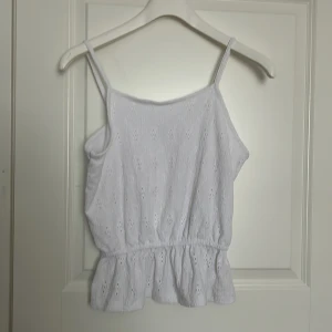 Vit topp från Kappahl - Säljer en söt vit topp från Kappahl med smala axelband och ett fint hålmönster. Toppen har en volangdetalj vid nederkanten och är tillverkad i polyester med lite stretch.