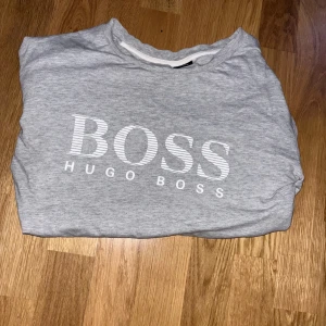Hugo Boss tröja  - Säljer en stilren grå tröja från Hugo Boss med deras ikoniska logga tryckt på framsidan. Tröjan har långa ärmar och en klassisk passform, perfekt för en avslappnad look.