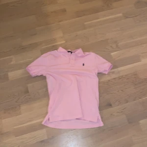 Rosa pikétröja från Ralph Lauren - Snygg rosa pikétröja från Ralph Lauren med klassisk passform. Tröjan har korta ärmar och en liten broderad logotyp på bröstet. Perfekt för en avslappnad och stilren look.