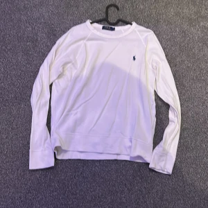 Vit långärmad tröja från Ralph Lauren - Säljer en stilren vit långärmad tröja från Ralph Lauren med det klassiska logotypen broderad på bröstet. Perfekt för en avslappnad och elegant look. Tröjan har en rund halsringning och är tillverkad i ett mjukt material.