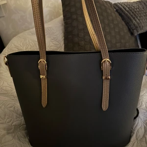 Grå/blå axelväska med bruna detaljer - Snygg blå/grå axelväska med bruna handtag och guldfärgade detaljer. Väskan har en dragkedja upptill och flera fack inuti för enkel organisering. Perfekt för att bära med sig det nödvändigaste med stil. I princip oanvänd. 