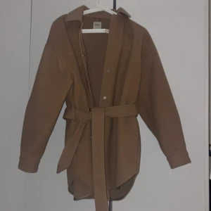 Beige kappa från ONLY - Beige kappa från ONLY med knappar, bälte i midjan och med fickor på sidorna. Perfekt till hösten och den är aldrig använd förutom nu vid uppvisning för annons. Märke är dock kvar från när jag tog bort fickor fram på bröstet.