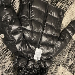 Svart pufferjacka från Moncler - Snygg svart pufferjacka från Moncler med glansig finish och dragkedja framtill. Jackan har en quiltad design och långa ärmar, perfekt för kyliga dagar. Den har också en praktisk huva och två fickor med dragkedja.