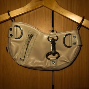 Beige axelväska med detaljer - Snygg beige axelväska i syntetmaterial med coola metallringar och en dragkedja framtill. Perfekt för att ge din outfit en edgy touch. Enkelt att fästa med valfritt axelband💕