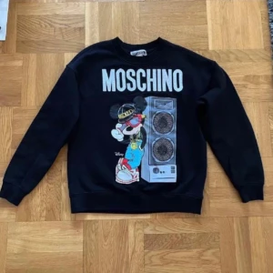 Svart sweatshirt från Moschino X H&M med Musse Pigg-tryck - Svart college tröja från Moschino med ett stort tryck av Musse Pigg och en högtalare på framsidan. Tröjan har långa ärmar och en rund halsringning. Har endast använt en gång.