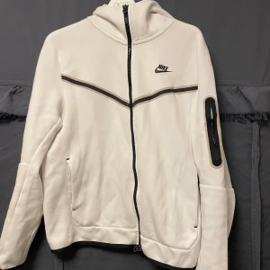 Nike tech fleeze - Säljer en vit hoodie från Nike med dragkedja och huva. Jackan har en svart randig detalj över bröstet och en ficka med dragkedja på ärmen. Perfekt för en sportig look.