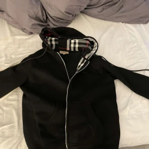 Burberry Kofta - Säljer en svart huvtröja från Burberry med dragkedja och klassiskt rutigt mönster i huvan. Tröjan har långa ärmar och en bekväm passform. Perfekt för en stilren och avslappnad look.
