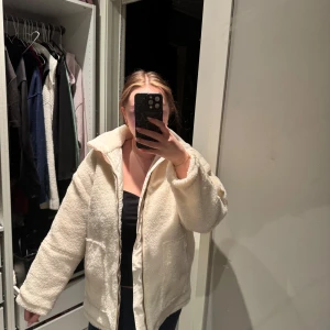 Vit teddyjacka med dragkedja - Mysig vit teddyjacka med dragkedja framtill. Jackan har en avslappnad passform och stora fickor. Perfekt för kyliga dagar när du vill hålla dig varm och bekväm.