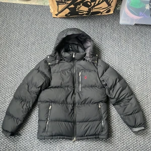 Svart dunjacka från Polo Ralph Lauren - Säljer en svart dunjacka från Polo Ralph Lauren med avtagbar huva och dragkedja. Jackan har en broderad röd logotyp på bröstet och praktiska fickor med dragkedjor. Perfekt för kyliga dagar! Använd i ca 3 månader så skicket är 10/10