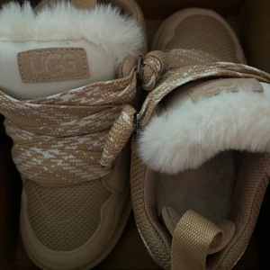 Beige sneakers från UGG - Snygga beige sneakers från UGG päls på insidan. Skorna har ett stickat mönster och snörning framtill. Perfekta för kyligare dagar med sin varma och bekväma design.