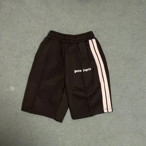 Svarta shorts från Palm Angels - Snygga svarta shorts från Palm Angels med vita ränder längs sidorna. De har en elastisk midja för extra komfort och stilren design med logotypen tryckt på framsidan. Perfekta för en avslappnad look.Storlek M men passar L