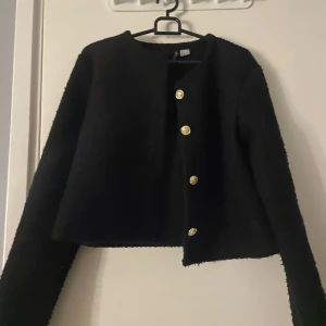 Svart kavaj från H&M - Snygg svart kavaj från H&M med guldknappar framtill. Den har en croppad design och långa ärmar, perfekt för en stilren look. Passar bra till både vardag och festliga tillfällen.