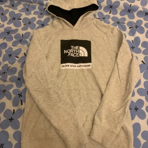  The North Face hoodie - Säljer en grå hoodie från The North Face med deras ikoniska logga och texten 'Never Stop Exploring' på framsidan. Tröjan har en bekväm passform med långa ärmar och en praktisk huva. Perfekt för kyliga dagar!