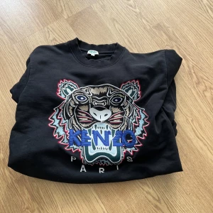 Svart sweatshirt från Kenzo med tigerbroderi - Säljer en svart sweatshirt från Kenzo med ett stort, färgglatt tigerbroderi på framsidan. Tröjan har långa ärmar och ribbade muddar vid ärmslut och nederkant. Perfekt för en stilren och trendig look. hyfsat använd men fortfarande i fint skick 