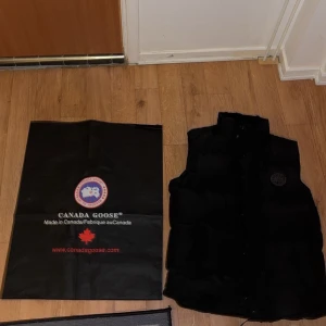 Svart dunväst från Canada Goose - Säljer en svart dunväst från Canada Goose. Västen har en hög krage och knappar framtill. Perfekt för kyligare dagar och har en stilren design med logotyp på bröstet.