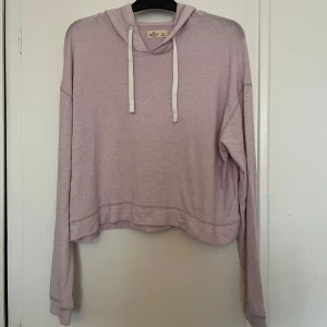 Ljusrosa hoodie från Hollister - Ljusrosa hoodie från Hollister. Den har en croppad design med långa ärmar och en justerbar huva med vita snören. Finns en liten fläck längst ner på framsidan men går att tvätta bort 🩷