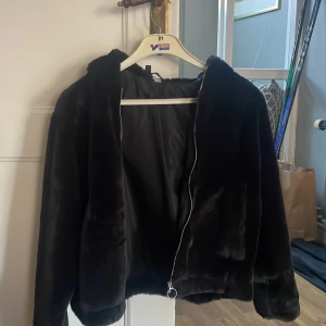 Svart pälsjacka från H&M Divided - Säljer en svart pälsjacka från H&M Divided med huva och dragkedja. Jackan har en mjuk och fluffig känsla, perfekt för kyligare dagar. Den har långa ärmar och två fickor framtill. En stilren och mysig jacka för höst och vinter.