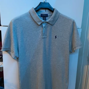 Grå pikétröja från Ralph Lauren - Säljer en stilren grå pikétröja från Ralph Lauren.  Skicket på tröjan är riktigt bra, knappt använd! Tröjan är storlek 18-20 i barnstorlek vilket motsvarar ungefär s i vanlig storlek 