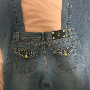 Blå jeans med nitar från Cartise - Snygga blå jeans från Cartise med unika detaljer som nitar och dekorativa knappar. Jeansen har en klassisk femficksdesign och en cool logotyp på baksidan. Perfekta för dig som vill sticka ut med stil.