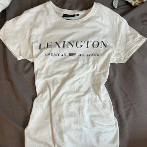 Vit t-shirt från Lexington - Säljer en vit t-shirt från Lexington med texten 'American Heritage' och en liten flagga på framsidan. T-shirten är kortärmad och tillverkad i ekologisk bomull. Perfekt för en avslappnad stil.