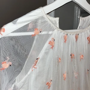 Vit klänning med flamingomönster - Charmig vit klänning med ett genomskinligt lager och broderade flamingor i rosa. Klänningen har långa ärmar och en luftig passform, perfekt för en somrig look. Den har en rund halsringning och ett lätt fall som ger en romantisk känsla.