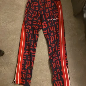 Mönstrade byxor från Palm Angels - Snygga byxor från Palm Angels med ett iögonfallande mönster i rött och blått. Byxorna har en sportig look med dragkedjor vid bensluten och ränder längs sidorna. Perfekta för dig som vill sticka ut!