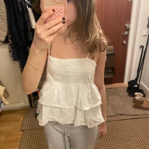 Vit ärmlös topp med spetsdetaljer - Säljer en söt vit ärmlös topp med smala axelband och spetsdetaljer. Toppen har en volangdesign som ger en luftig och somrig känsla. Perfekt för varma dagar!