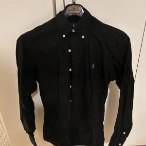 Svart skjorta från Ralph Lauren - Snygg svart skjorta från Ralph Lauren med klassisk krage och knappar framtill. Den har en broderad logga i lila på bröstet. Perfekt för en stilren look.