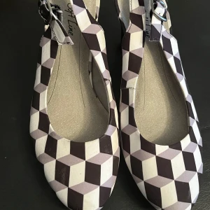 Mönstrade ballerinaskor från Soft Line - Snygga ballerinaskor från Soft Line med ett unikt geometriskt mönster i svart och vit. Skorna har en justerbar rem med spänne för bättre passform. Perfekta för att ge en stilfull touch till din outfit.