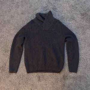 Grå stickad tröja från Fred Perry - Säljer en stilren grå stickad tröja från Fred Perry i 100% lammull. Tröjan har en hög krage och långa ärmar med snygga armbågslappar i mörkare ton. Perfekt för kyliga dagar!
